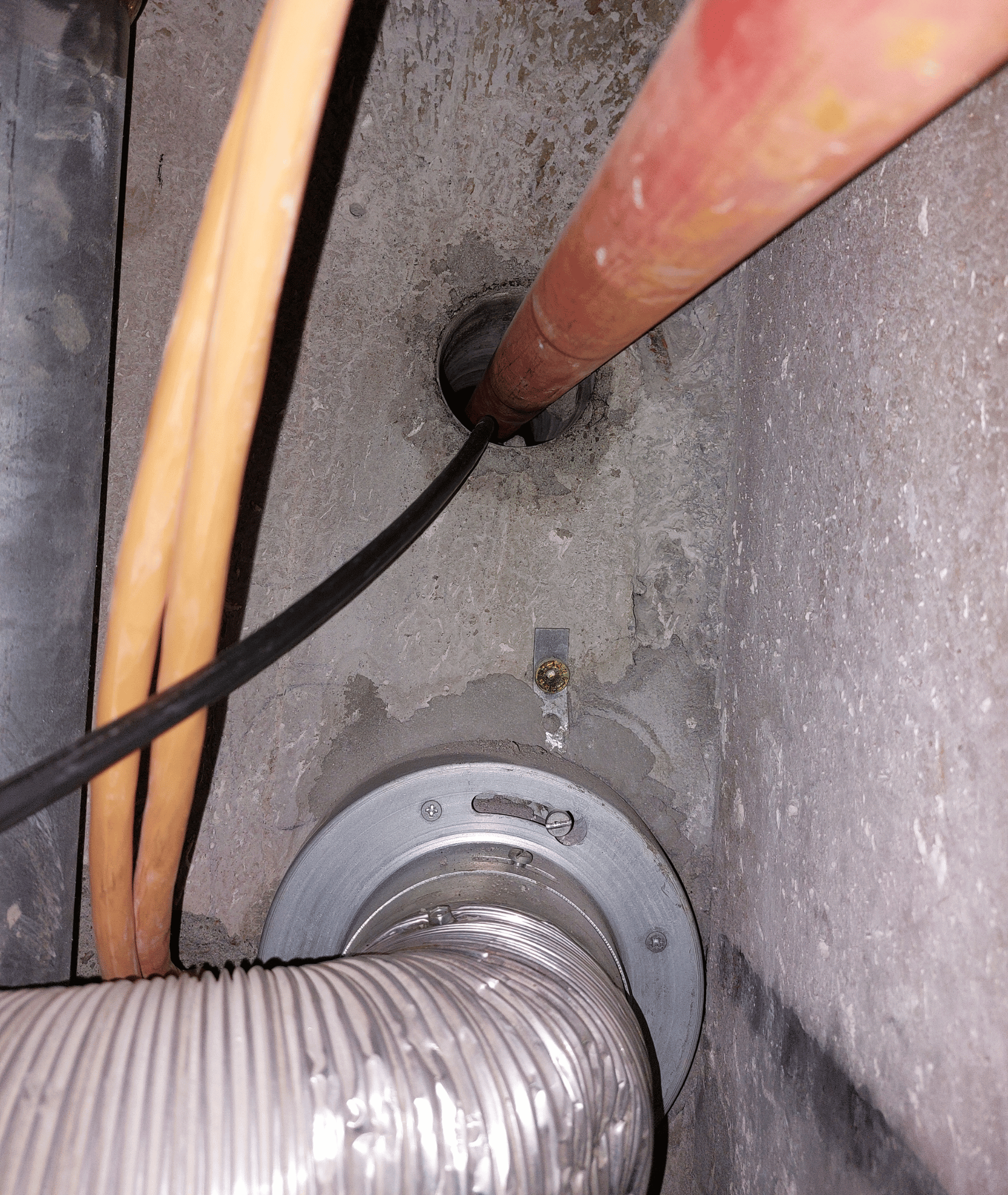 Aufnahme einer Wand. Im unteren Bereich ist eine Brandschutzklappe, die an einem flexiblen Schlauch angeschlossen ist, zu sehen. Oberhalb der Klappe ist eine Rohrdurchführung durch eine offene Kernbohrung zu sehen. Durch diese Kernbohrung ist zusätzlich ein elektrisches Kabel geführt