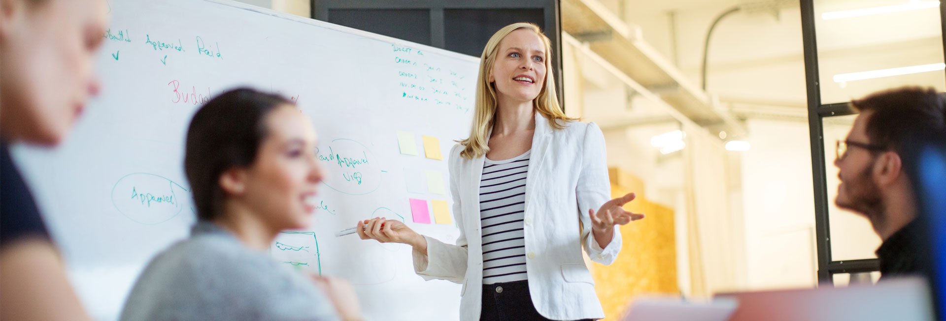 Frau spricht vor einer Gruppe mit Whiteboard im Hintergrund.