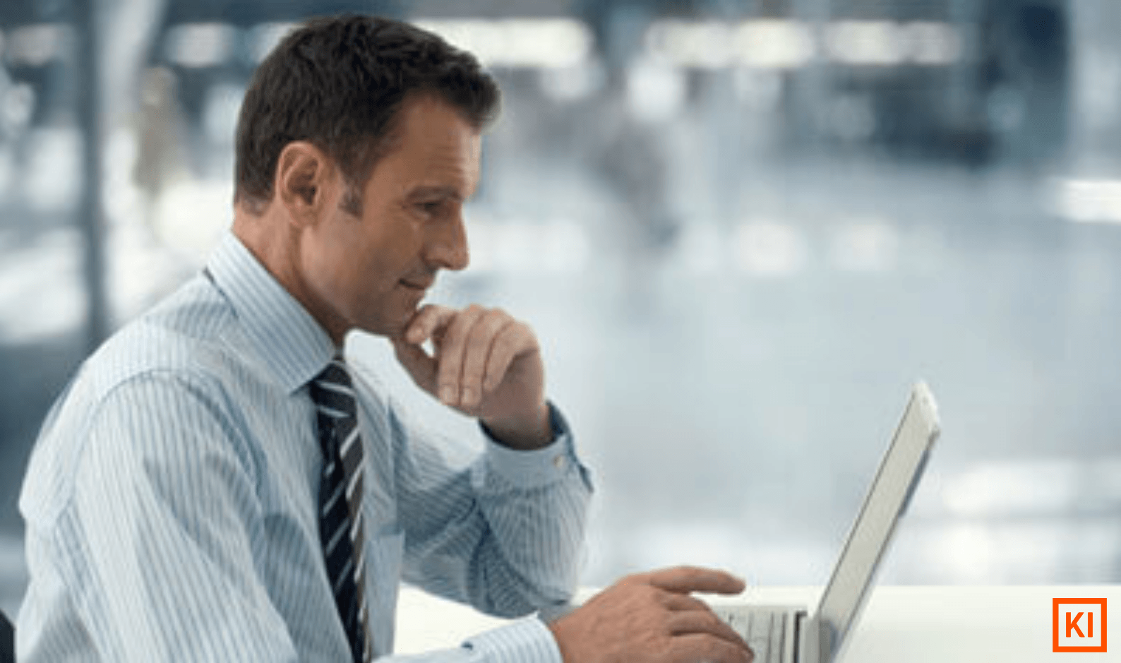 Mann in Hemd und Krawatte arbeitet konzentriert an einem Laptop in einem modernen, hellen Büro.