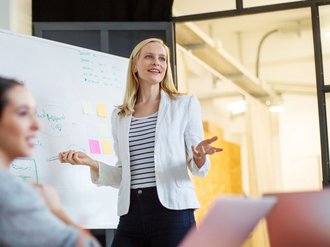 Frau spricht vor einer Gruppe mit Whiteboard im Hintergrund.